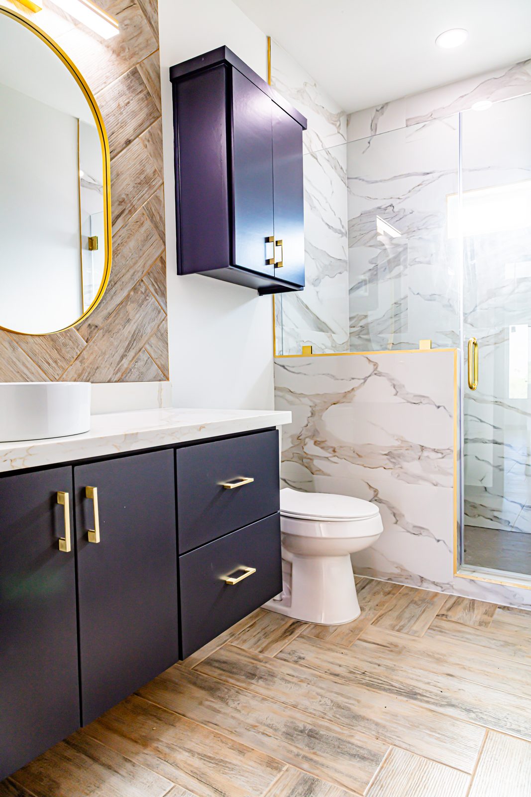 Bathroom with navy vanity, gold accents, marble shower surround, and round gold mirror