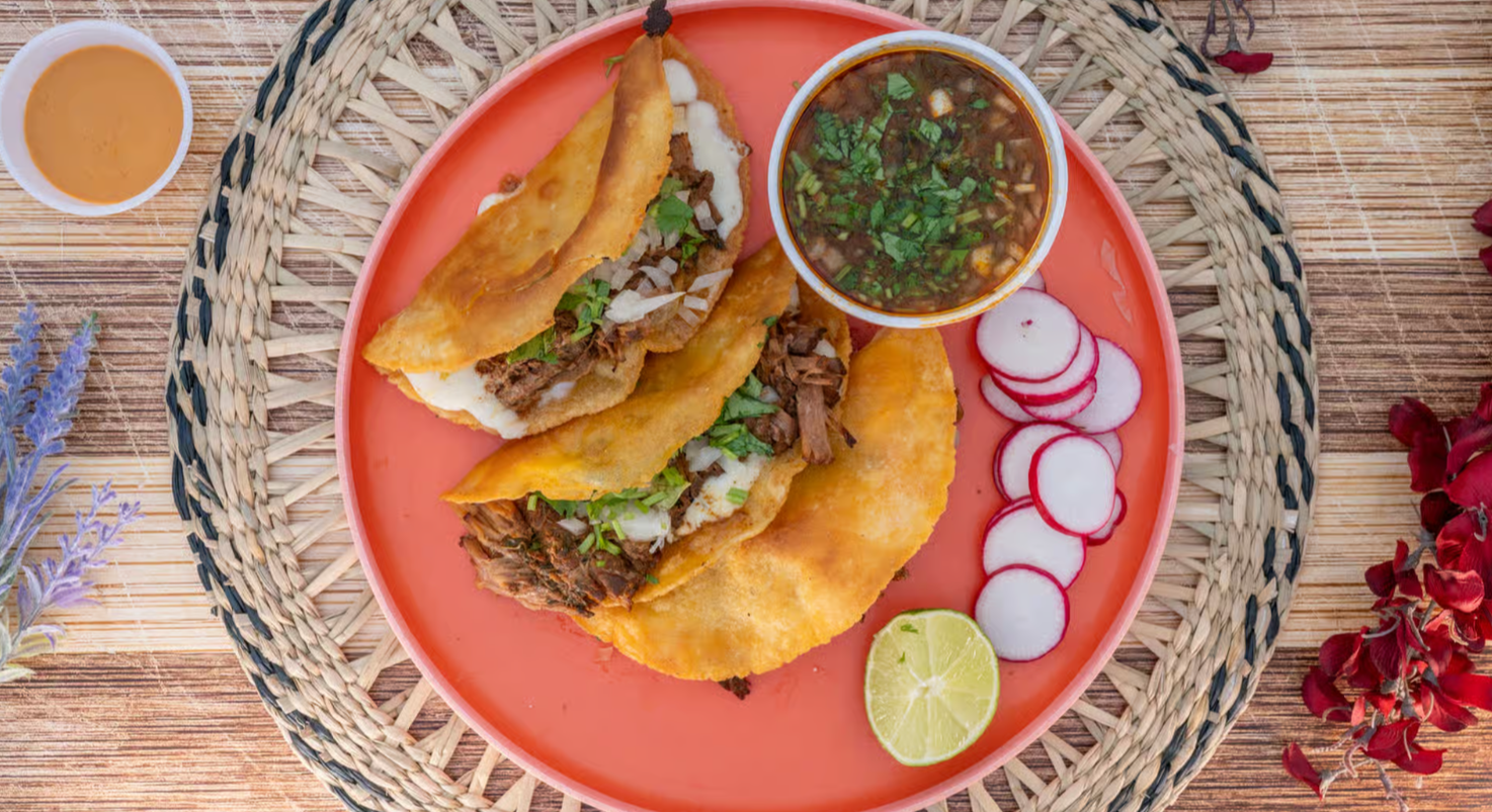 Authentic Mexican Quesabirria tacos with melted Oaxaca cheese, slow-braised beef birria, crispy golden tortillas, garnished with fresh cilantro, diced white onion, and lime wedges, served alongside a steaming bowl of rich red consomme dipping broth, captured in moody dark food photography