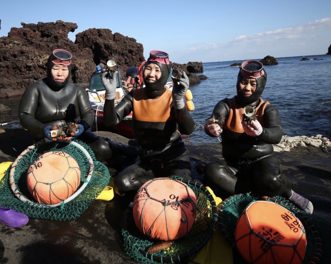 Traditional Haenyeo sea women divers in their iconic gear standing on the rocky volcanic coast of Jeju with crashing ocean waves behind them.