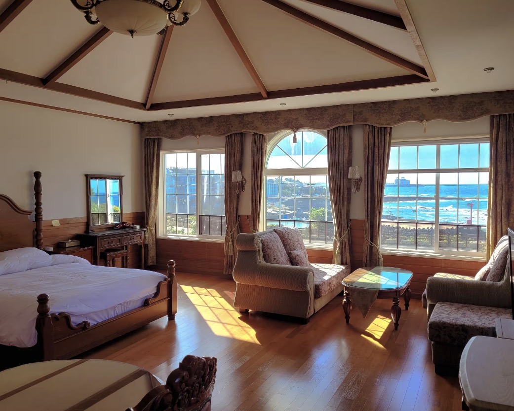 A bright, airy hotel room with a private balcony showing a stunning view of the blue Jeju ocean at sunrise.
