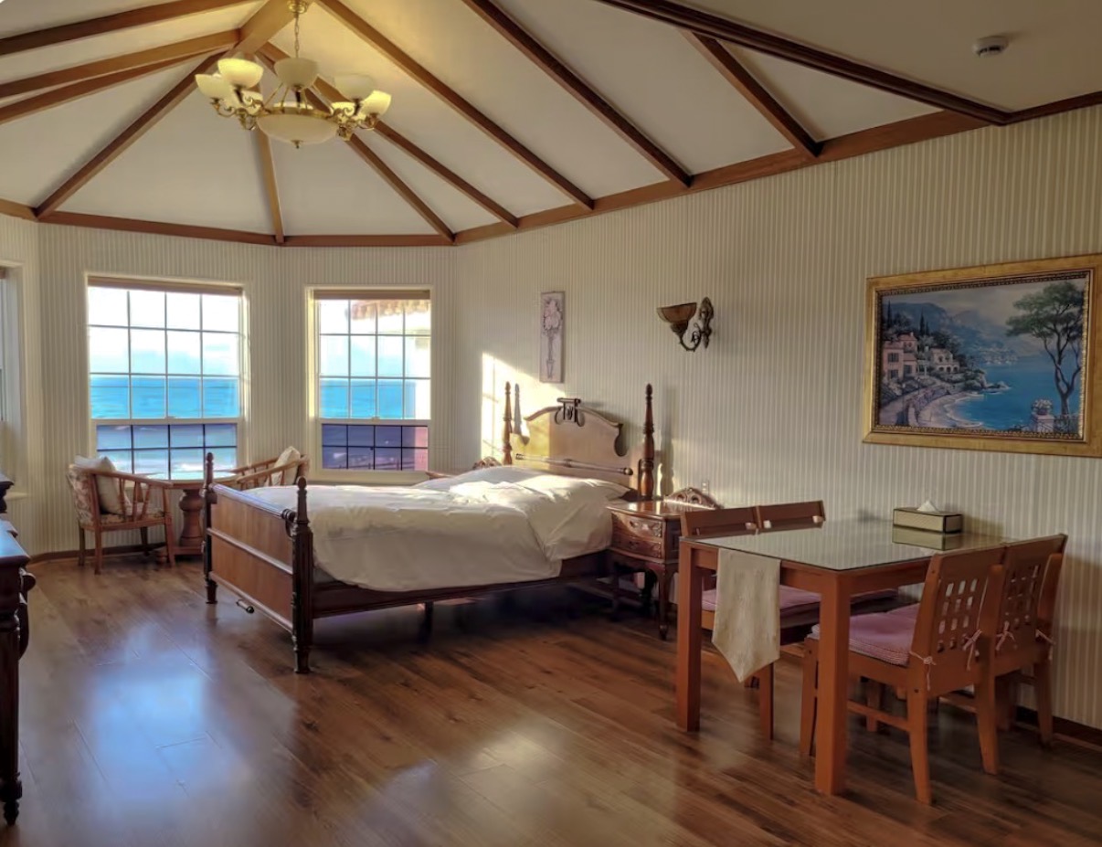 A bright, airy hotel room with a private balcony showing a stunning view of the blue Jeju ocean at sunrise.