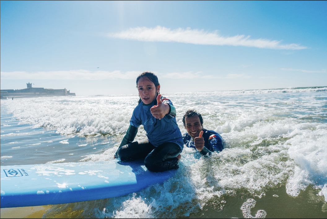 Fullscreen documentary photograph of adaptive surfing session with surfer and volunteer instructor riding a wave together at Carcavelos beach, Portugal