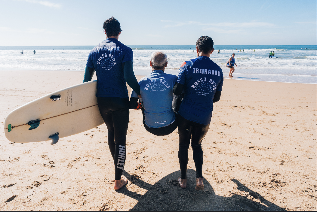 Retrato de um aluno de surf adaptado a sorrir após uma sessão em Carcavelos, com o mar ao fundo
